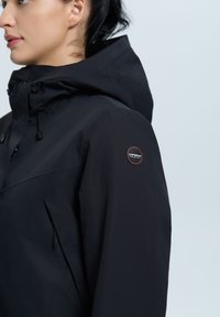 Svart vattentät jacka med hög krage, justerbar huva och ficka med dragkedja på bröstet. Har en logotyp-patch på vänster axel.