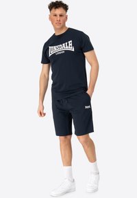 Tamnoplava pamučna majica s printom "Lonsdale London", uparen s odgovarajućim šortsevima i bijelim tenisicama. Model stoji s rukama u džepovima.