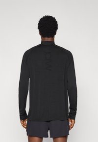 Μακρυμάνικο μαύρο αθλητικό τοπ με ψηλό γιακά, ραγκλάν μανίκια και διακριτική λεπτομέρεια λογότυπου στο πίσω μέρος, συνδυασμένο με σκούρο σορτς.