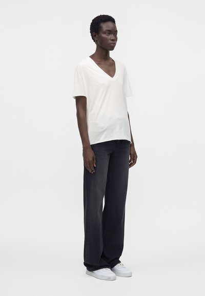Γυναίκα όρθια με λευκό μπλουζάκι V-neck, φαρδιά μαύρα τζιν και λευκά αθλητικά παπούτσια σε απλό λευκό φόντο.