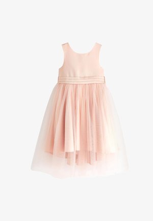Mädchen mit langen dunklen Haaren, das ein ärmelloses, hellrosa Kleid trägt, steht vor pastellfarbenen Ballons und hellrosa Vorhängen.