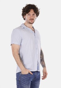 Camicia azzurra chiara a maniche corte con colletto, dal tessuto liscio e con una leggera lucentezza; indossata con jeans in denim scuro e tatuaggi visibili.