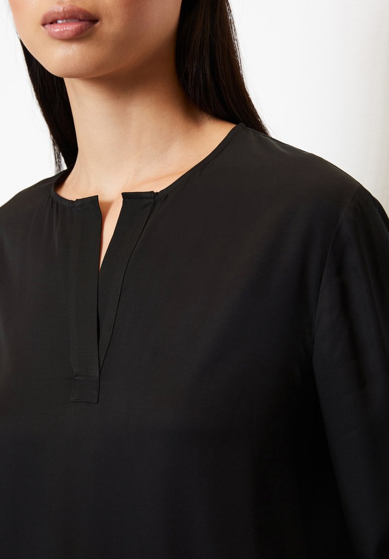 Czarna bluzka z okrągłym dekoltem i częściowym zapięciem, wykonana z lekkiego materiału o gładkiej fakturze. Brak widocznych wzorów lub akcentów.