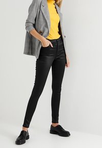 Šedý kostkovaný blazer s velkými knoflíky, žlutý žebrovaný rolák, vysoké černé kalhoty z umělé kůže a černé šněrovací boty.