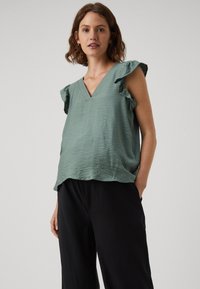 Lichtgroene mouwloze top met een V-hals en gerimpelde schouders, gemaakt van textuurstof; gedragen met zwarte wijde broek.