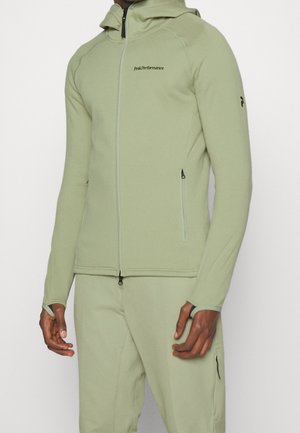 Homme portant une veste à capuche zippée vert clair et un pantalon assorti, la veste comporte des fermetures éclair à l'avant et sur les manches ainsi que des trous pour les pouces.