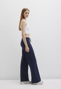 Navy wide-leg jeans made of denim, paired with a white cropped tank top. The model wears silver athletic shoes, standing against a plain background.