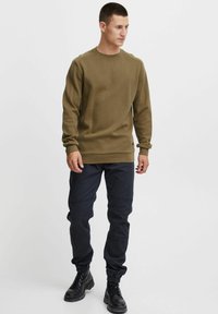 Sweatshirt vert olive avec des manches longues, en tissu texturé, col rond, associé à un pantalon de jogging bleu marine et des bottes noires à lacets.