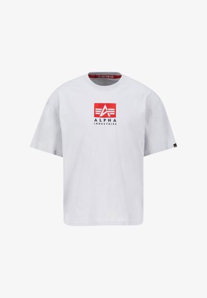 Fehér pamut póló kerek nyakkal, elöl piros négyzet alakú logóval, amelyen fehér és fekete szöveg olvasható: "ALPHA INDUSTRIES". Rövid ujjú.