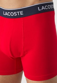 Sarkanas kokvilnas bokseršorti ar tumši zilu jostas vietu, kas rotāta ar "LACOSTE" logo un kontrastējošām baltām svītrām. Gluda tekstūra, pieguļoša forma.