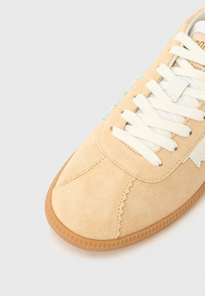 Μπεζ αθλητικό παπούτσι από σουέτ με λευκά κορδόνια, που διαθέτει υφή σόλας από καουτσούκ και διακοσμητικό μοτίβο στην πλευρά, αναδεικνύοντας έναν χαλαρό σχεδιασμό.