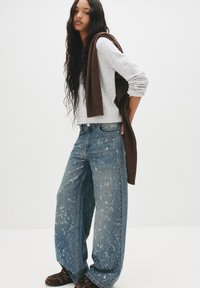Pulover gri deschis, blugi denim albaștri clasați cu model de stropi, și o eșarfă maro aruncată pe umăr. Pantofi cu model maro și negru.