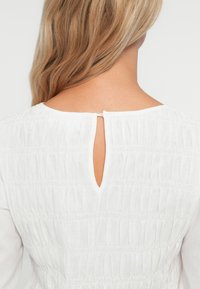 Hvid bluse med smock-design på bagsiden, med en nøglehulåbning og et blødt, tekstureret materiale.
