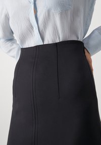 Svart skräddarsydd kjol med smal passform, som har två främre veck, en slät yta och hög midja. Bärs med en ljusblå knapp-up skjorta.