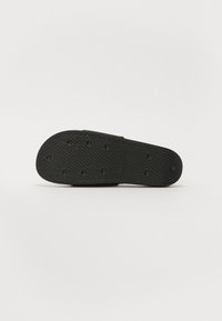 Black rubber sandal sole with a textured pattern and circular grip spots. Flat, open design with a low profile for easy wear.