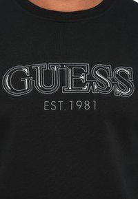 Fekete anyag, rajta a "GUESS" felirat fehér körvonalas betűkkel, fölötte pedig kisebb fehér szöveggel: "EST. 1981".