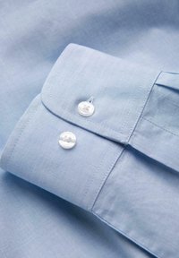 Poignet de chemise bleu clair avec deux boutons blancs et des coutures détaillées sur un fond en tissu lisse.