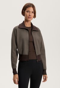 Brown and cream patterned zip-up jacket with a high collar, ribbed cuffs, and fitted design, paired with a brown top and dark pants.