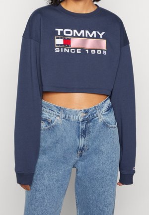 Osoba nosząca granatową krótką bluzę z napisem i logo „TOMMY JEANS SINCE 1985”, zestawioną z jasnoniebieskimi, wysokim stanem jeansami.