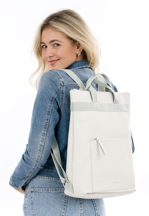 Jauna moteris dėvi džinsinę striukę, ant peties nešiodama baltą stačiakampį kuprinę su šviesiai pilkomis sagtimis, šypsosi į kamerą.