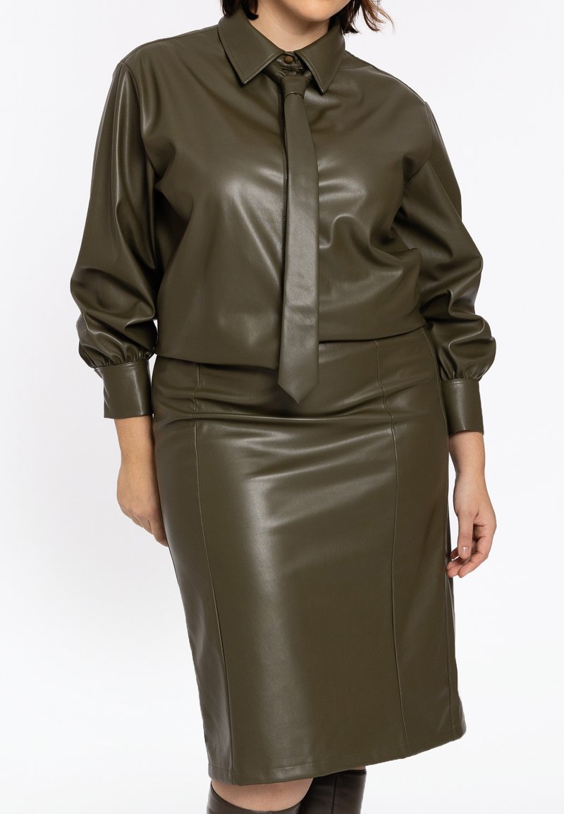 Kvinde iført en olivengrøn bluse i imiteret læder med matchende slips og pencilnederdel, stående med hænder afslappet ved siderne.