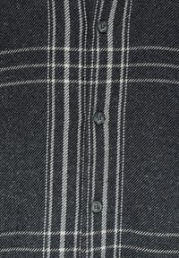 Tissu gris foncé avec motif à carreaux blancs et trois boutons noirs visibles alignés verticalement sur un vêtement.