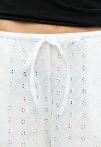 Κοντινή λήψη λευκού υφασμάτινου παντελονιού με μικρές λουλουδένιες τρύπες και φαρδιά κορδόνια δένονται στη μέση, σε συνδυασμό με μαύρο τοπ.