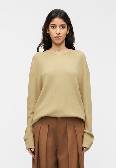 Pull en maille côtelée couleur kaki clair avec une coupe décontractée, épaules tombantes et manches longues, associé à un pantalon plissé marron.
