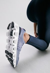Grauer Sportschuh mit Obermaterial aus Netzgewebe und Gummisohle, der ein markantes mehrschichtiges Design und einen schwarzen strukturierten Grip an der Außensohle aufweist.