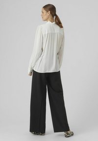 Witte blouse met lange mouwen en geplooide details aan de achterkant, gecombineerd met ruime zwarte broeken en zwart-wit gestreepte schoenen.