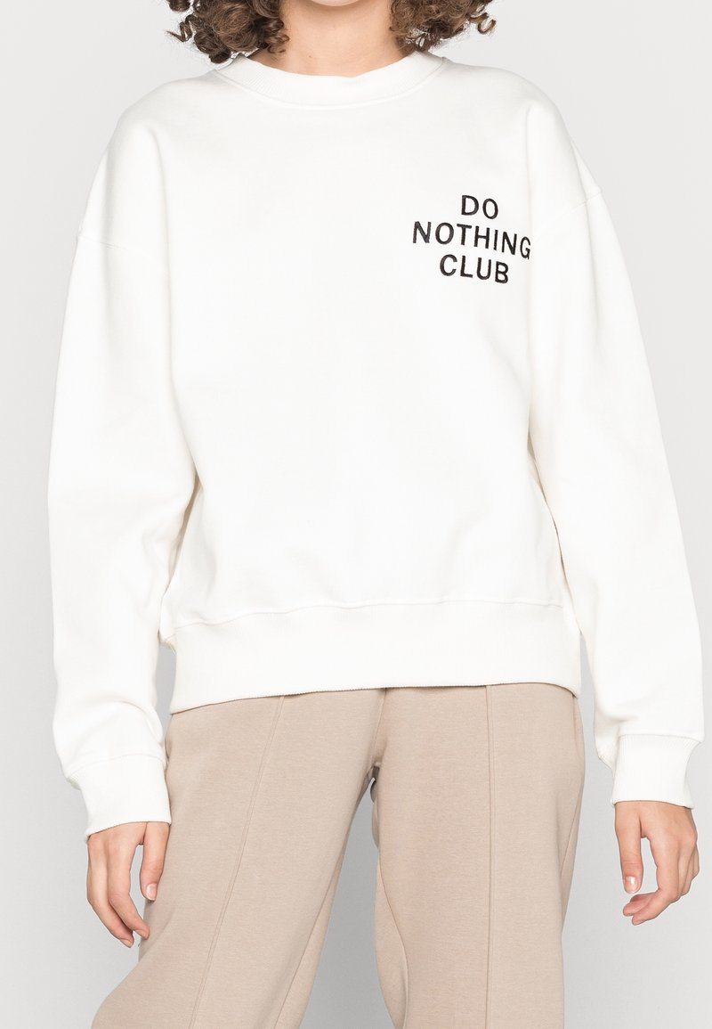 Bílá mikina s žebrovanými manžetami a spodním lemem, na přední straně s černým nápisem "DO NOTHING CLUB". Měkký materiál s volným střihem.