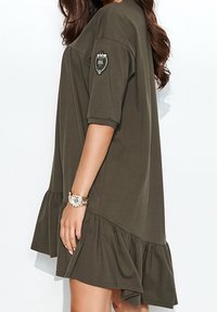 Vestido corto de manga corta en verde oliva con dobladillo acampanado, que presenta un parche decorativo en la manga y está hecho de una tela ligera.