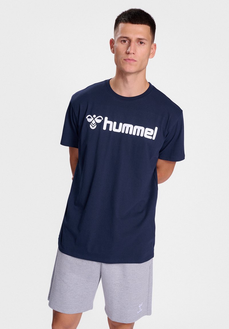 Tamsiai mėlynas medvilninis marškinėliai su trumpomis rankovėmis, su ryškiu baltu "hummel" logotipu ant krūtinės. Dėvimi su pilkais šortais.