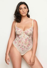 Nainen, jolla on keskipitkä tumma hius, pukeutuneena kukkakuvioiseen pitsiseen beigeen alusvaatebodyn, seisoo tasaisella vaalealla taustalla.