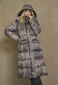 Donna che indossa un lungo cappotto imbottito a stampa leopardata con cappuccio, zip frontale e tasche laterali su uno sfondo beige uniforme.