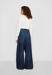 Kvinne med krøllete hår iført en hvit langermet topp og høyhåndte vide mørkeblå denimjeans vendt bort fra kamera.