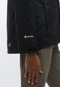 Μανίκι μαύρου μπουφάν Gore-Tex με ρύθμιση βέλκρο στο μανίκι, με έντονο λογότυπο. Το χέρι είναι τοποθετημένο πάνω σε γκρι παντελόνι.