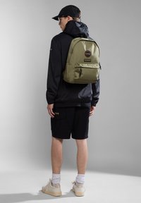 Zaino verde oliva con tasca frontale, dettagli neri e logo rotondo; indossato sopra una giacca nera e pantaloni corti, con una forma compatta e rettangolare.