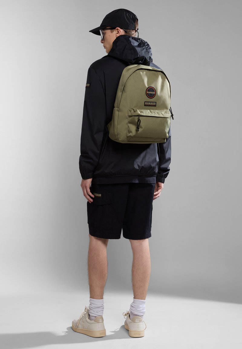 Zaino verde oliva con tasca frontale, dettagli neri e logo rotondo; indossato sopra una giacca nera e pantaloni corti, con una forma compatta e rettangolare.