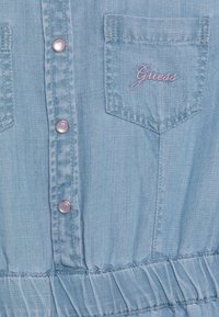 Vaaleansininen chambray-mekko, jossa nappikiinnitys edessä, kaksi rintataskua ja röyhelöyksityiskohta vyötäröllä. Taskussa kirjailtu logo.