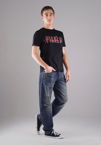 Schwarzes Baumwoll-T-Shirt mit rot-grauer Grafikschrift, kombiniert mit blauen, ausgewaschenen Jeans und schwarzen Canvas-Sneakern mit weißen Sohlen.