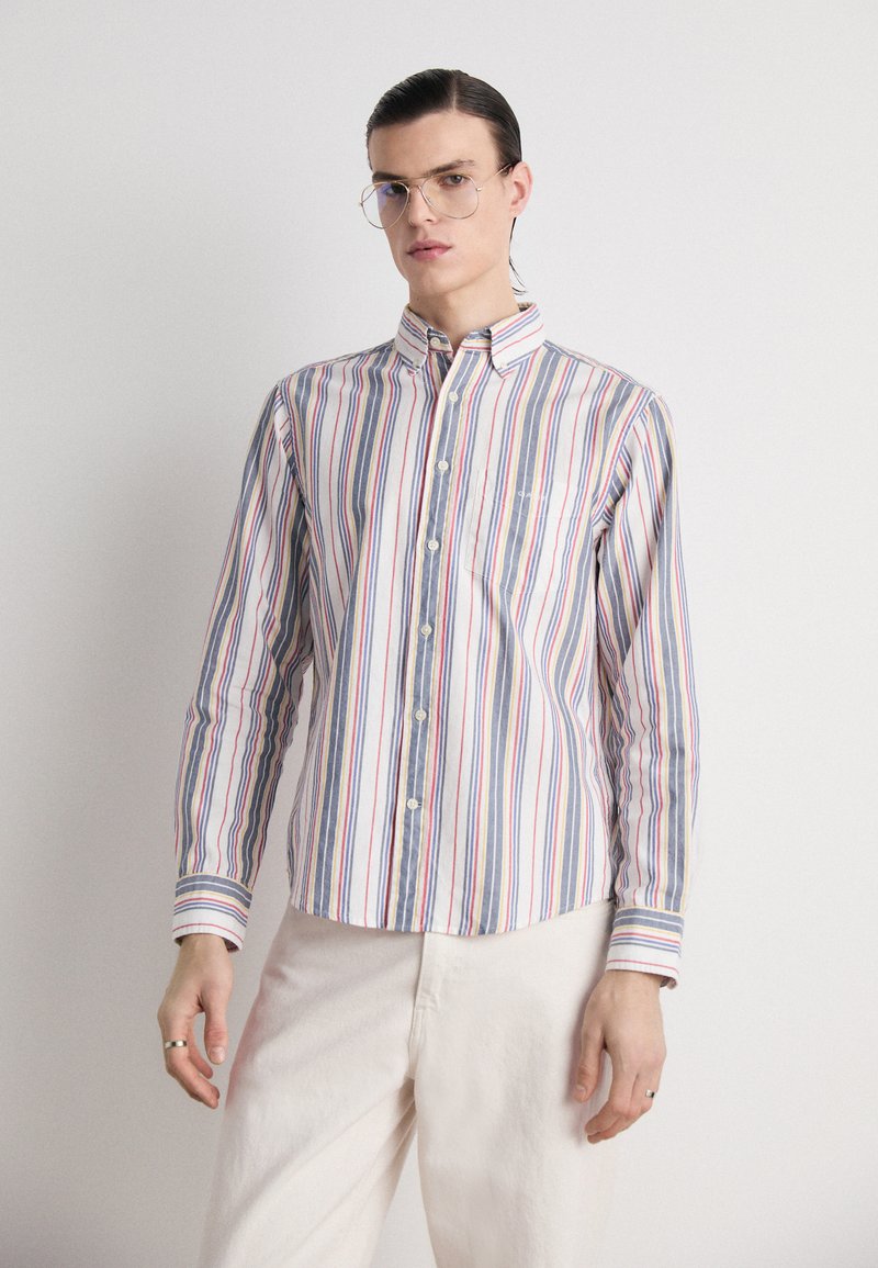 Camisa de botones a rayas con rayas azules, rojas y amarillas sobre un fondo blanco, con un bolsillo en el pecho y cuello clásico.