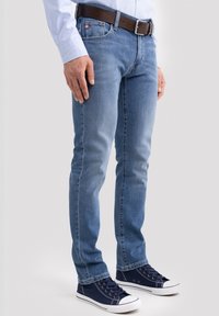 Vyras dėvi šviesiai mėlynus džinsus, rudos spalvos diržą, šviesiai mėlyną marškinius ir tamsiai mėlynas sportbačius, stovintis prie paprasto šviesaus fono.