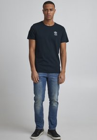 Marineblå bomulds t-shirt med rund hals og et lille grafisk design på brystet, parret med slidte blå jeans og sorte sneakers.