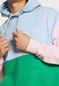 Felpa con cappuccio in blocchi di colore pastello blu, rosa e verde. Presenta una tasca frontale e testo ricamato bianco sul petto. Materiale in cotone.