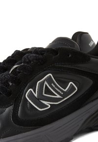 Melnās ādas un zamša sporta kurpes ar teksturētām akcentiem, biezām auklām un izteiktu baltu logotipu sānos. Gumijas zole, noapaļota forma.