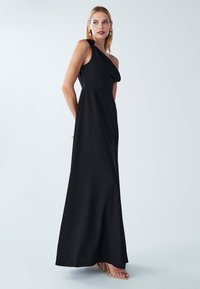 Czarna maxi sukienka na jedno ramię z miękkiego materiału, z dopasowanym gorsetem i zwiewną spódnicą, ozdobiona skręconym detalem na ramieniu.