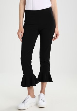 Woman wearing black cropped pants with flared ruffle hems and white sneakers, standing against a plain light background.