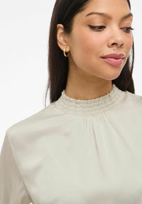 Vrouw met lang donker haar, draagt gouden creolen en een crèmekleurige blouse met hoge hals en geplooide details, kijkt opzij.