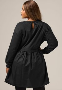 Musta mekko pitkillä hihoilla, tiukalla vartalolla ja laskostetulla hameella. Avainaukko-yksityiskohta takana. Pehmeä, teksturoitu kangas.